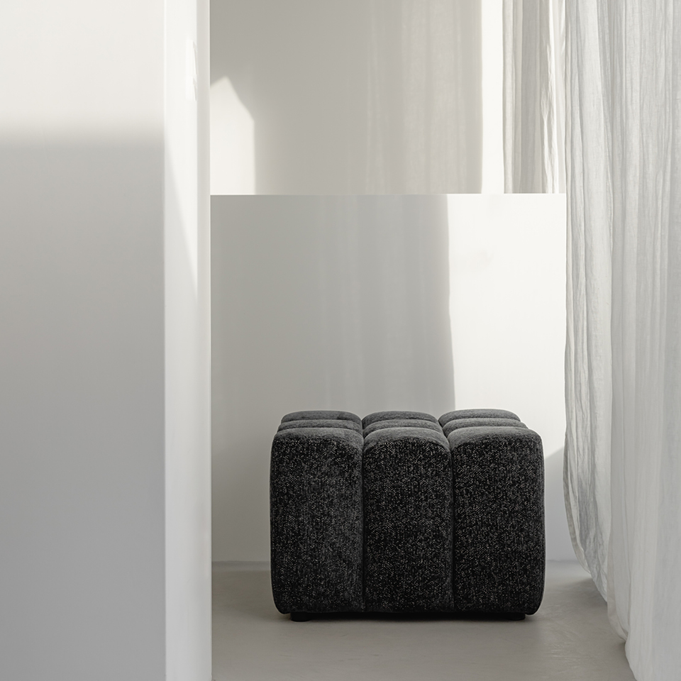 Minimalistische Szene mit einem dunklen, gepolsterten Hocker, der in einer hellen Nische zwischen weißen Wänden und leichten, durchscheinenden Vorhängen steht.