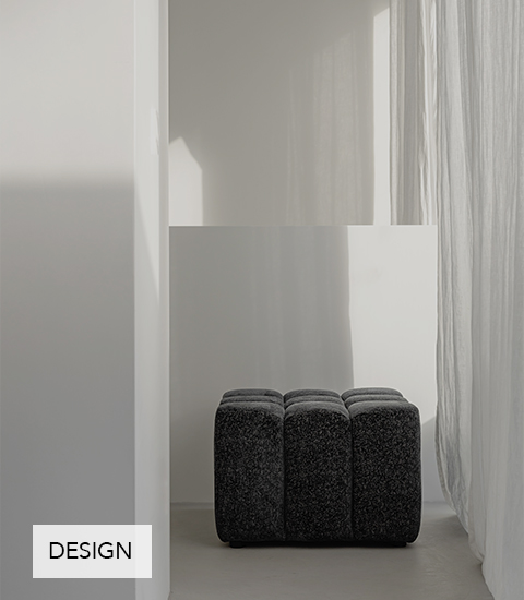 Minimalistische Szene mit einem dunklen, gepolsterten Hocker, der in einer hellen Nische zwischen weißen Wänden und leichten, durchscheinenden Vorhängen steht.