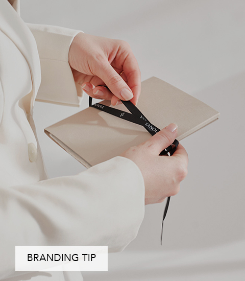 Eine Person im weißen Blazer hält ein beige-farbenes Notizbuch in den Händen und bindet ein schwarzes Satinband darum.