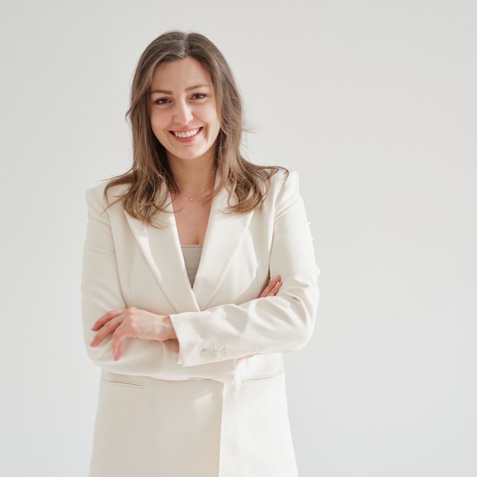 Tanja Grill, Gründerin und Creative Director bei TANOLARA im  eleganten cremefarbenen Hosenanzug steht vor hellem Hintergrund, verschränkt die Arme und lächelt selbstbewusst in die Kamera.