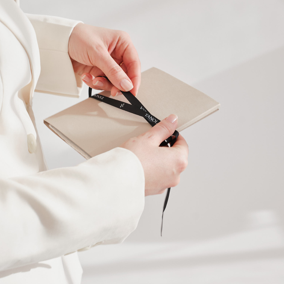 Eine Person im weißen Blazer hält ein beige-farbenes Notizbuch in den Händen und bindet ein schwarzes Satinband darum.