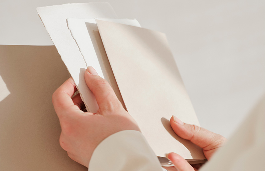 Eine Person hält mehrere Papierproben in neutralen Beige- und Weißtönen in den Händen, darunter auch handgeschöpftes Papier mit gerissenen Kanten.
