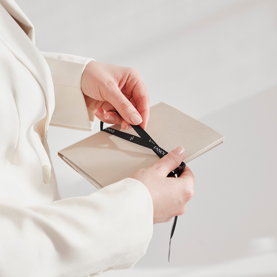 Eine Person im weißen Blazer hält ein beige-farbenes Notizbuch in den Händen und bindet ein schwarzes Satinband darum.