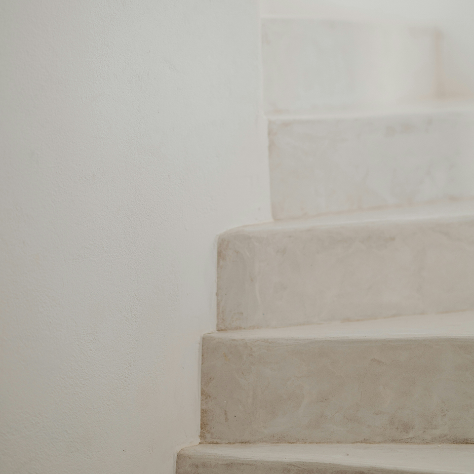 Eine schlichte, helle Steintreppe mit sanft gerundeten Kanten, die minimalistisch in einen weißen Putzwandhintergrund übergeht.