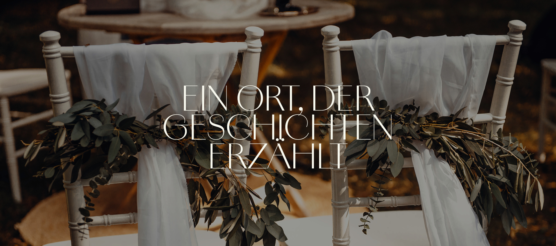 Zwei festlich dekorierte Stühle im Vintage-Stil, verziert mit weißen Stoffbahnen und grünen Zweigen. Darüber liegt der Text: „Ein Ort, der Geschichten erzählt“.